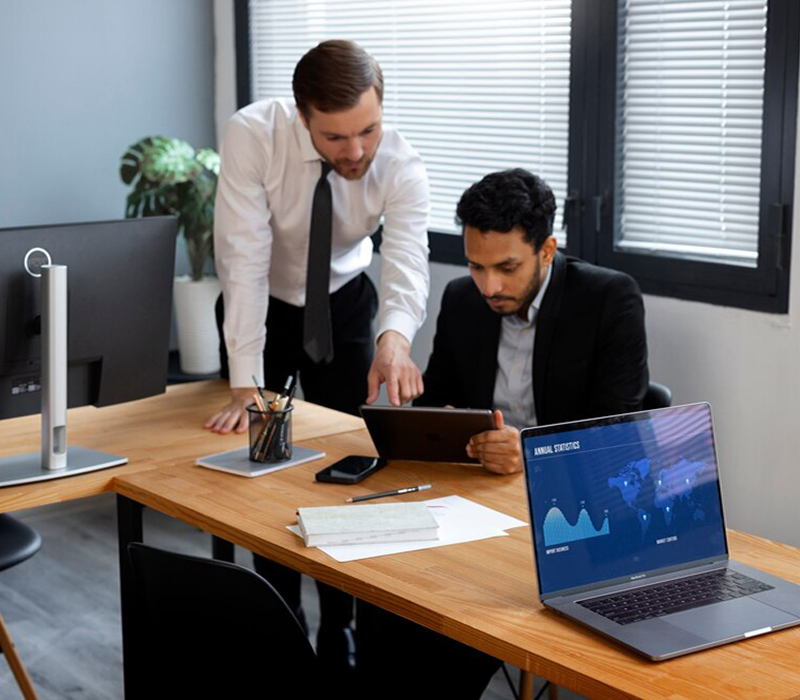 Two professionals reviewing data on a tablet with charts and annual statistics displayed on a laptop.